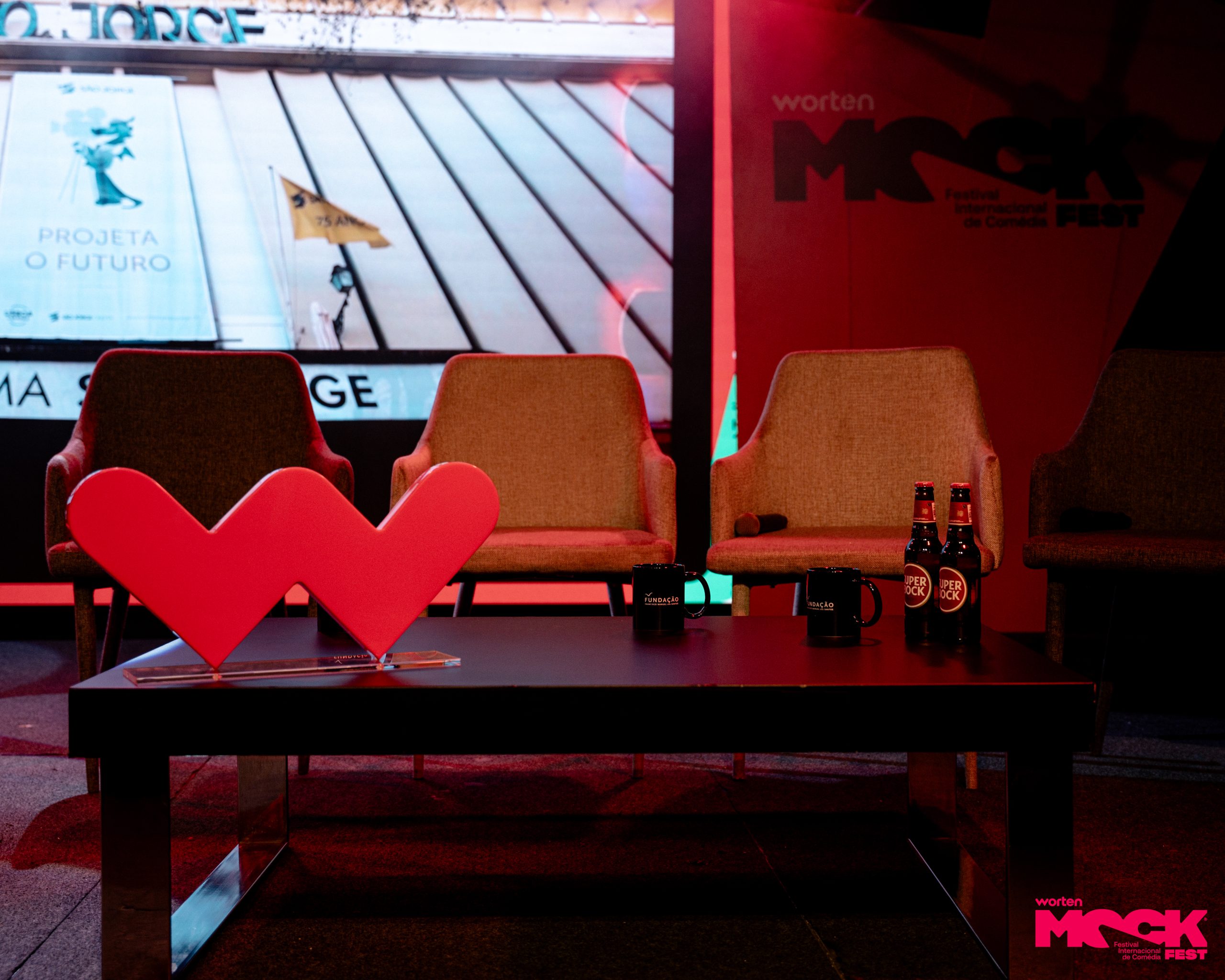 Mesa de palco com o logo vermelho da Worten em destaque, duas canecas pretas da Fundação Francisco Manuel dos Santos, duas garrafas de cerveja Super Bock e três cadeiras vazias ao fundo. No ecrã, vê-se parte de uma faixa com o texto “PROJETA O FUTURO”. No canto direito, o logotipo do Worten Mock Fest.