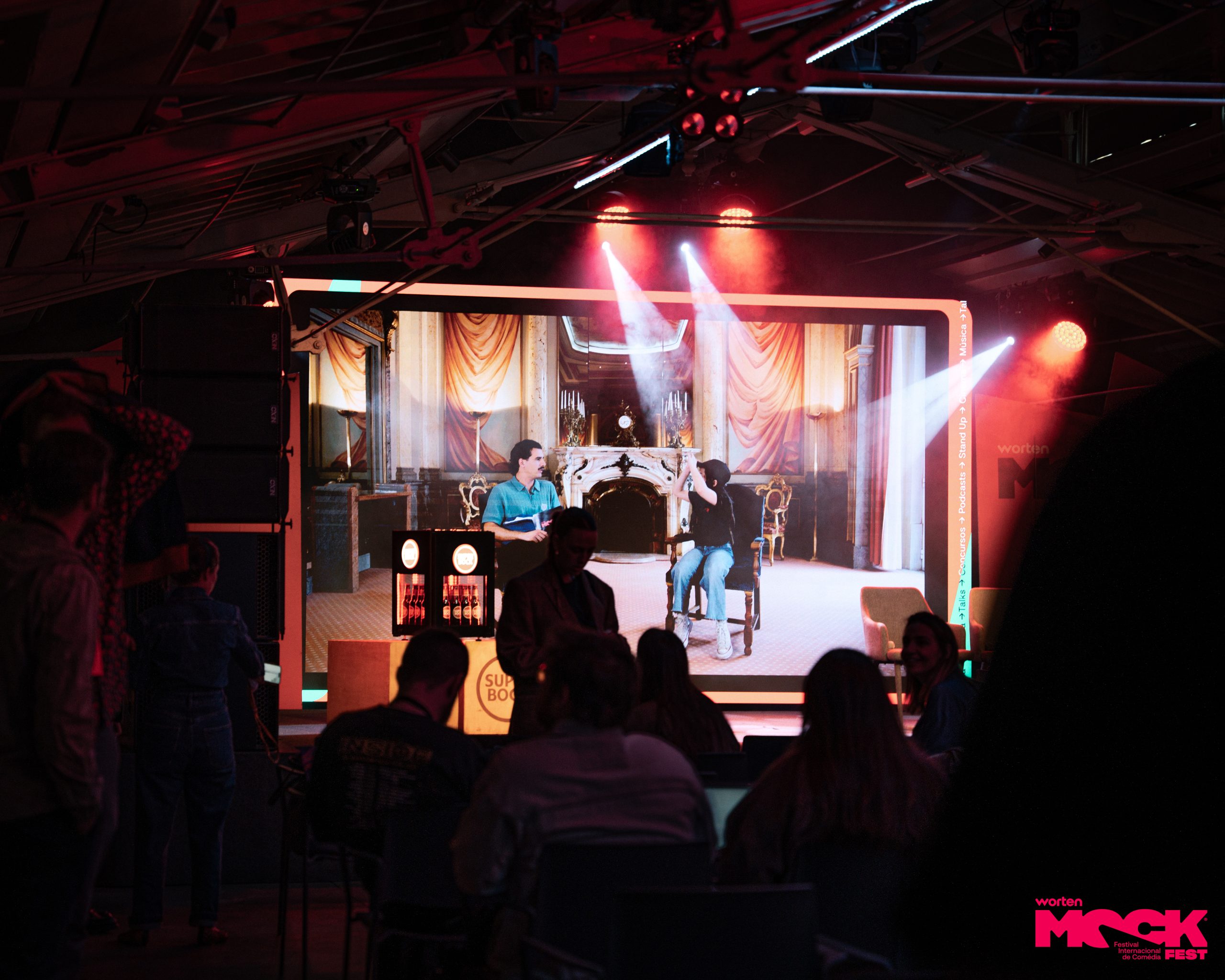Plateia assistindo apresentação humorística com cenário projetado no palco do Worten Mock Fest em Lisboa, com luzes e atmosfera de festival de comédia ao vivo.