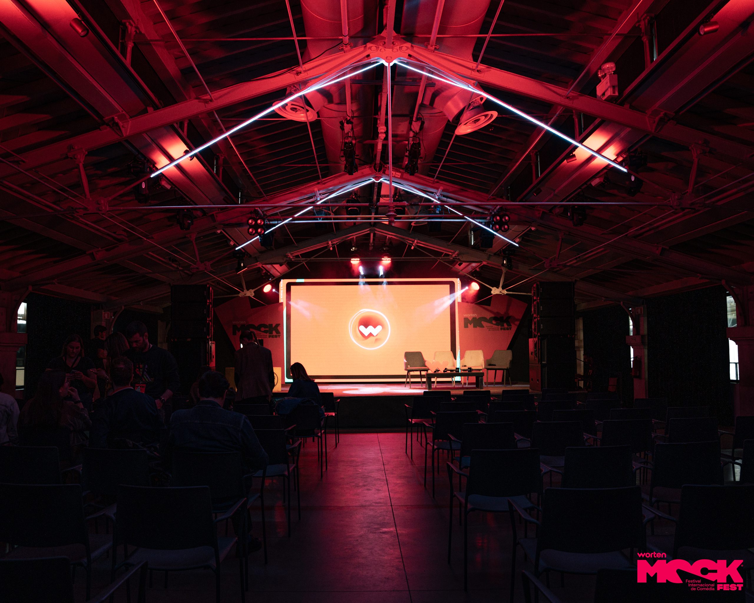 Palco principal do Worten Mock Fest iluminado com luzes vermelhas, cadeiras vazias e tela grande ao fundo com o logotipo do evento.