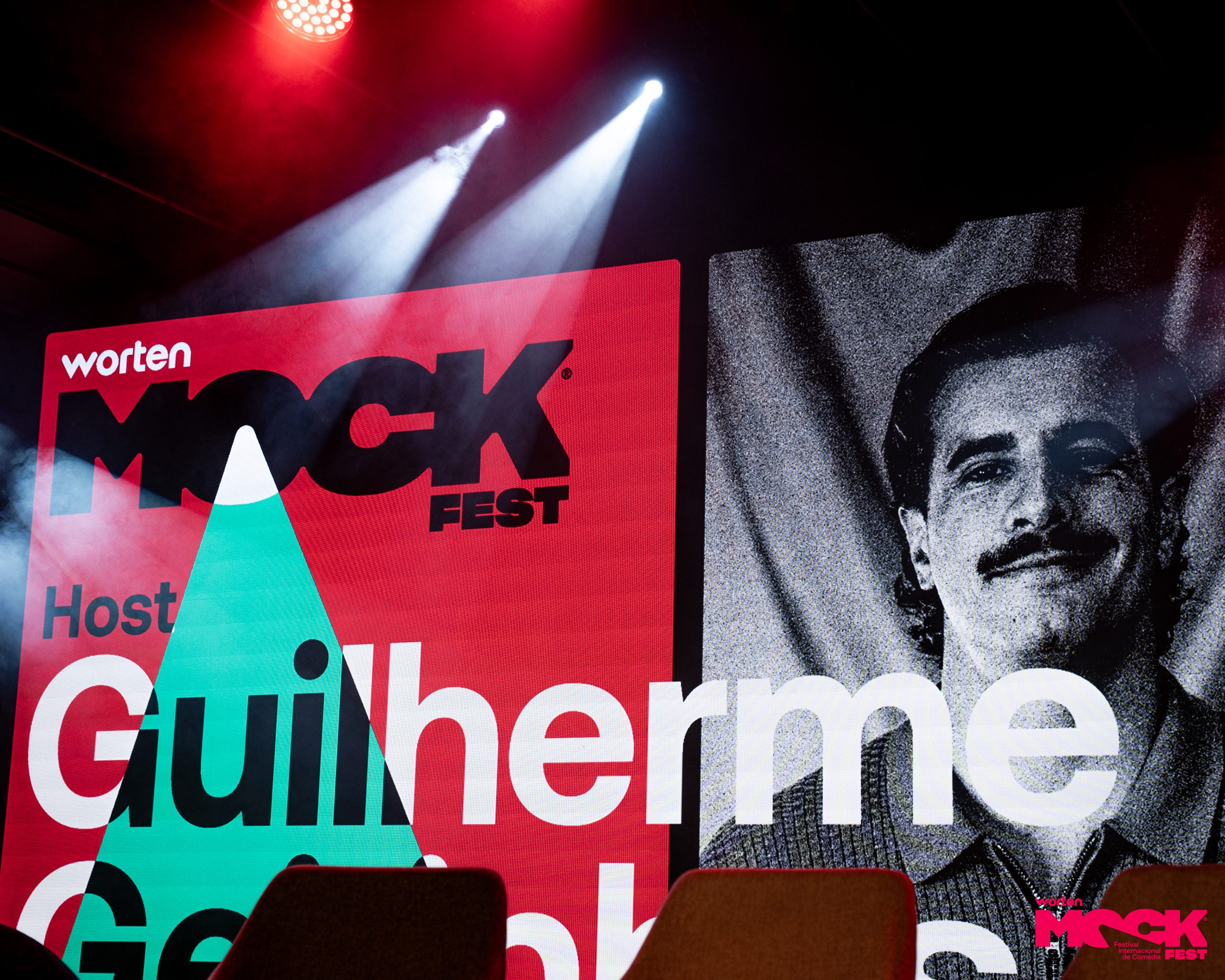 Ecrã iluminado no Worten Mock Fest com o nome do apresentador Guilherme e o logotipo do festival, com foco em luzes e imagem em preto e branco do host.