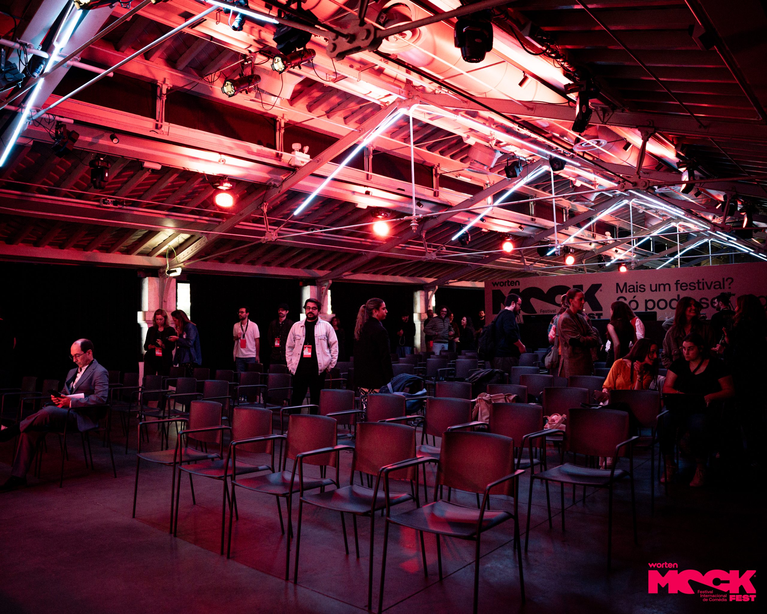 Sala de eventos do Mock Fest com cadeiras vazias em destaque, algumas pessoas conversando ao fundo e iluminação de festival, criando um ambiente descontraído antes do início das apresentações.