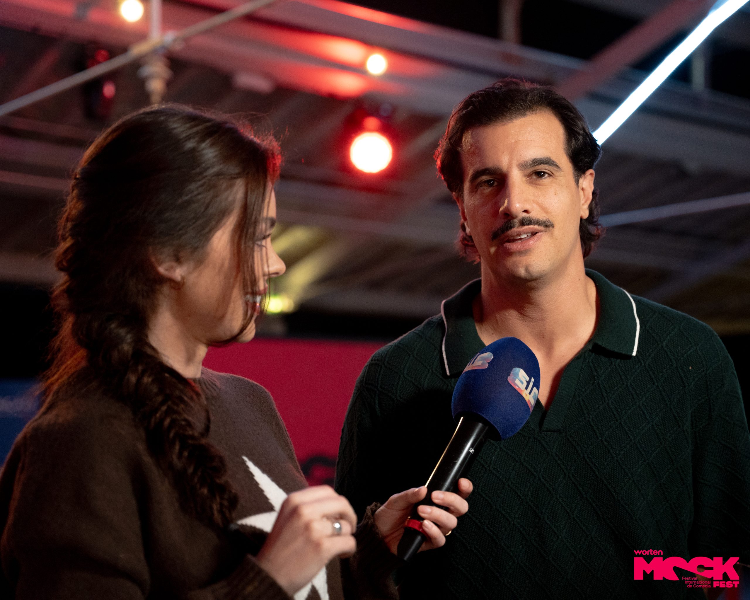 Entrevista no Worten Mock Fest. Repórter da SIC faz perguntas para um dos participantes do evento, que responde com expressão séria. Cena descontraída, típica de cobertura de festival de comédia.