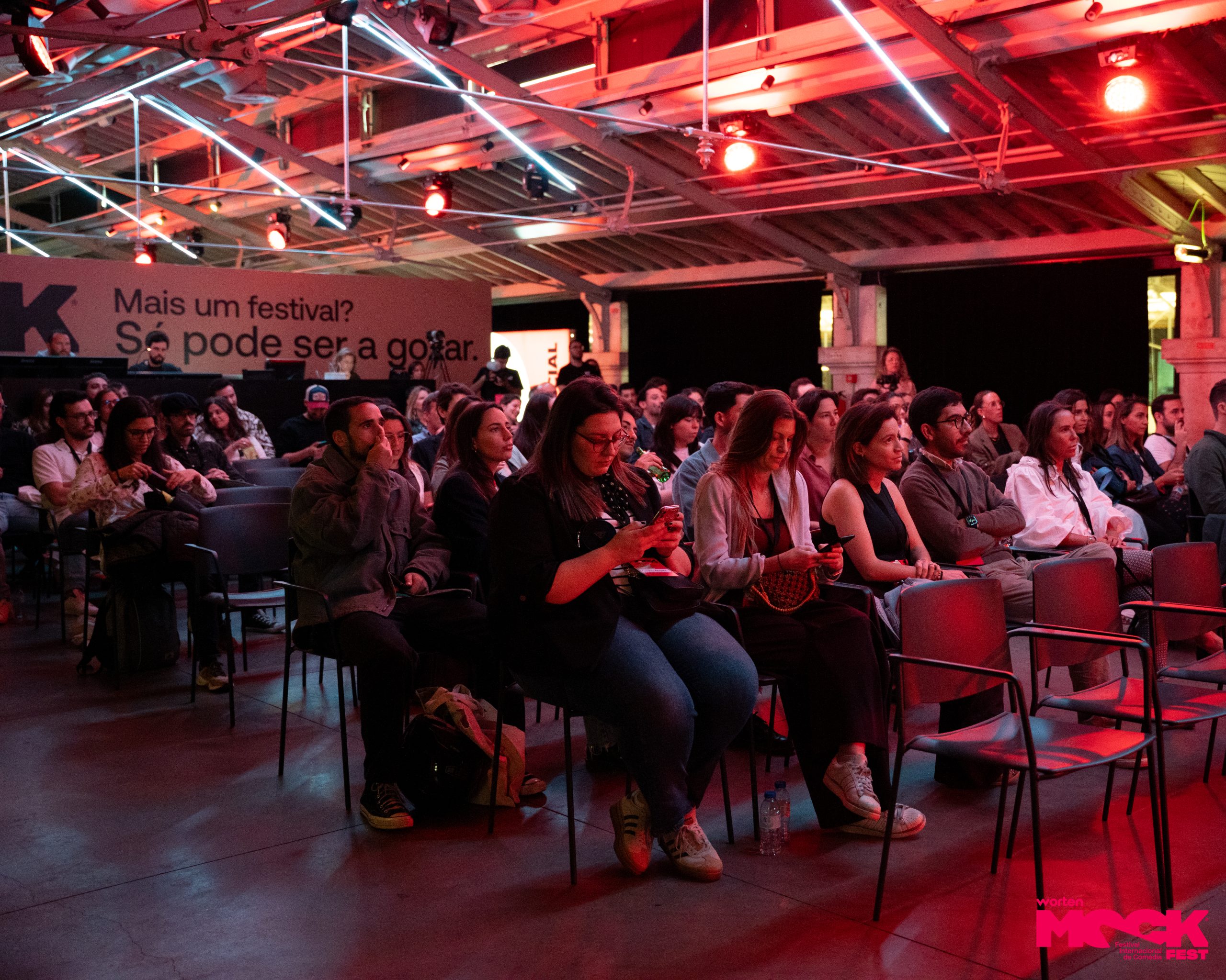 Plateia do Worten Mock Fest com pessoas sentadas e atentas ao evento, em ambiente iluminado com luzes vermelhas e o slogan “Só pode ser a gozar” visível ao fundo.
