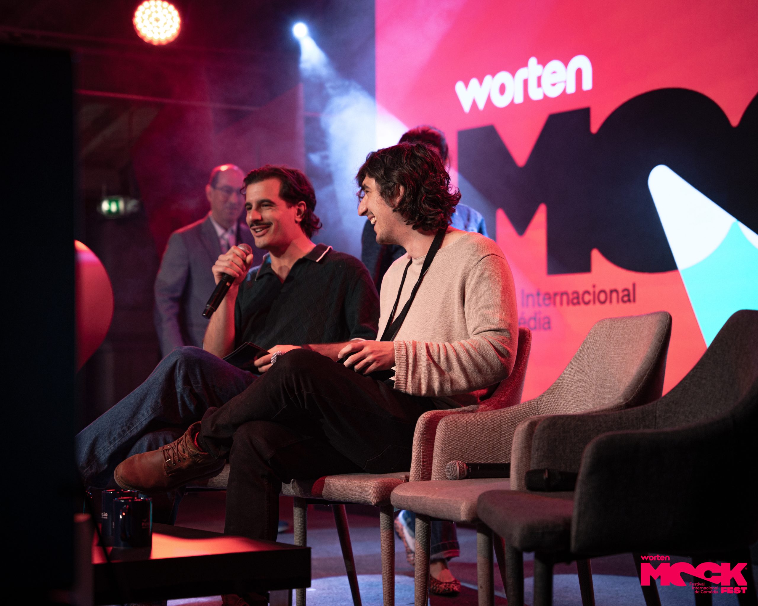 Comediantes no palco do Worten Mock Fest em um painel animado, sorrindo e conversando diante de uma plateia, com um grande logotipo vermelho em destaque à frente.