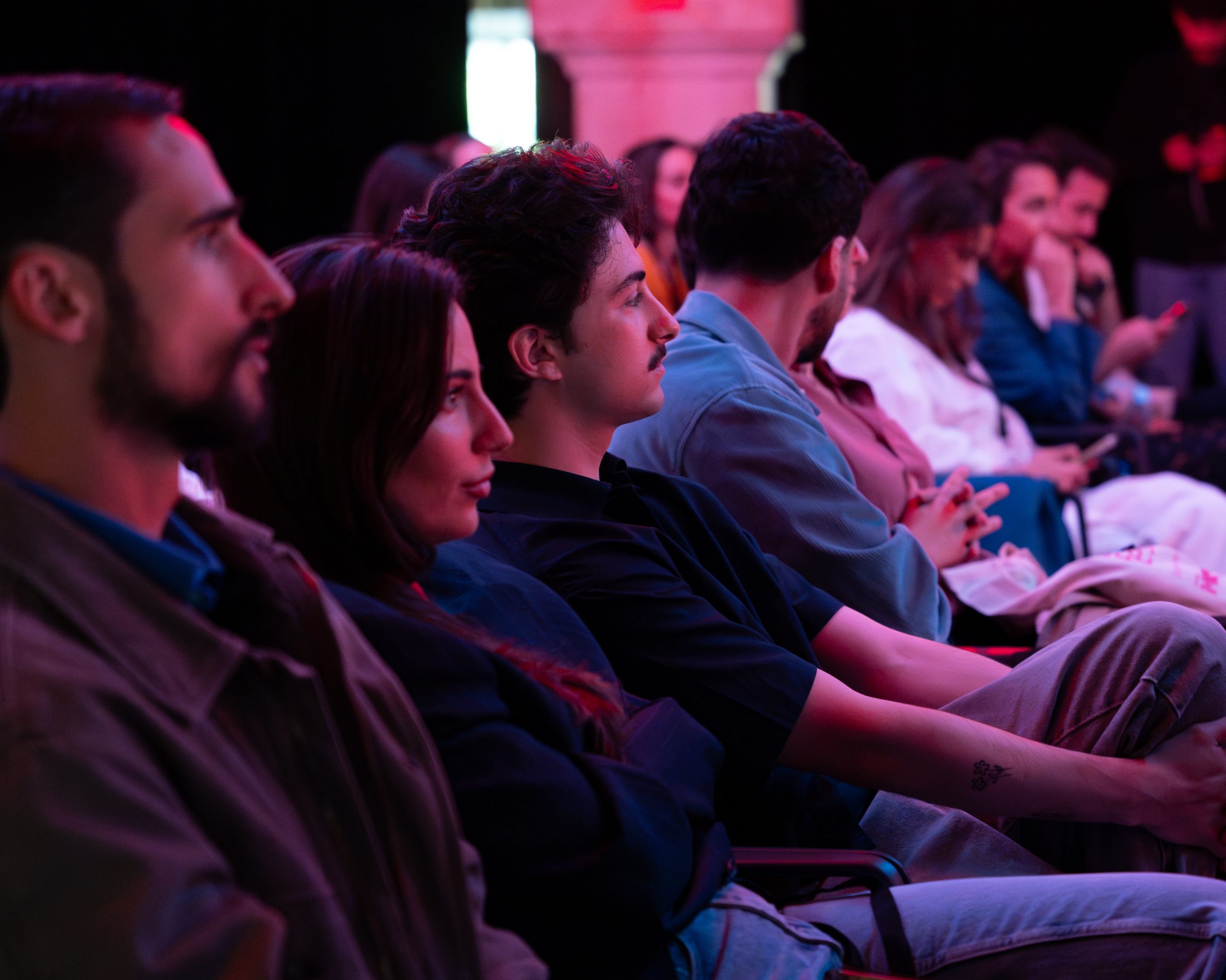 Público atento assistindo a uma apresentação durante o Mock Fest, com destaque para um homem careca e de barba na plateia.