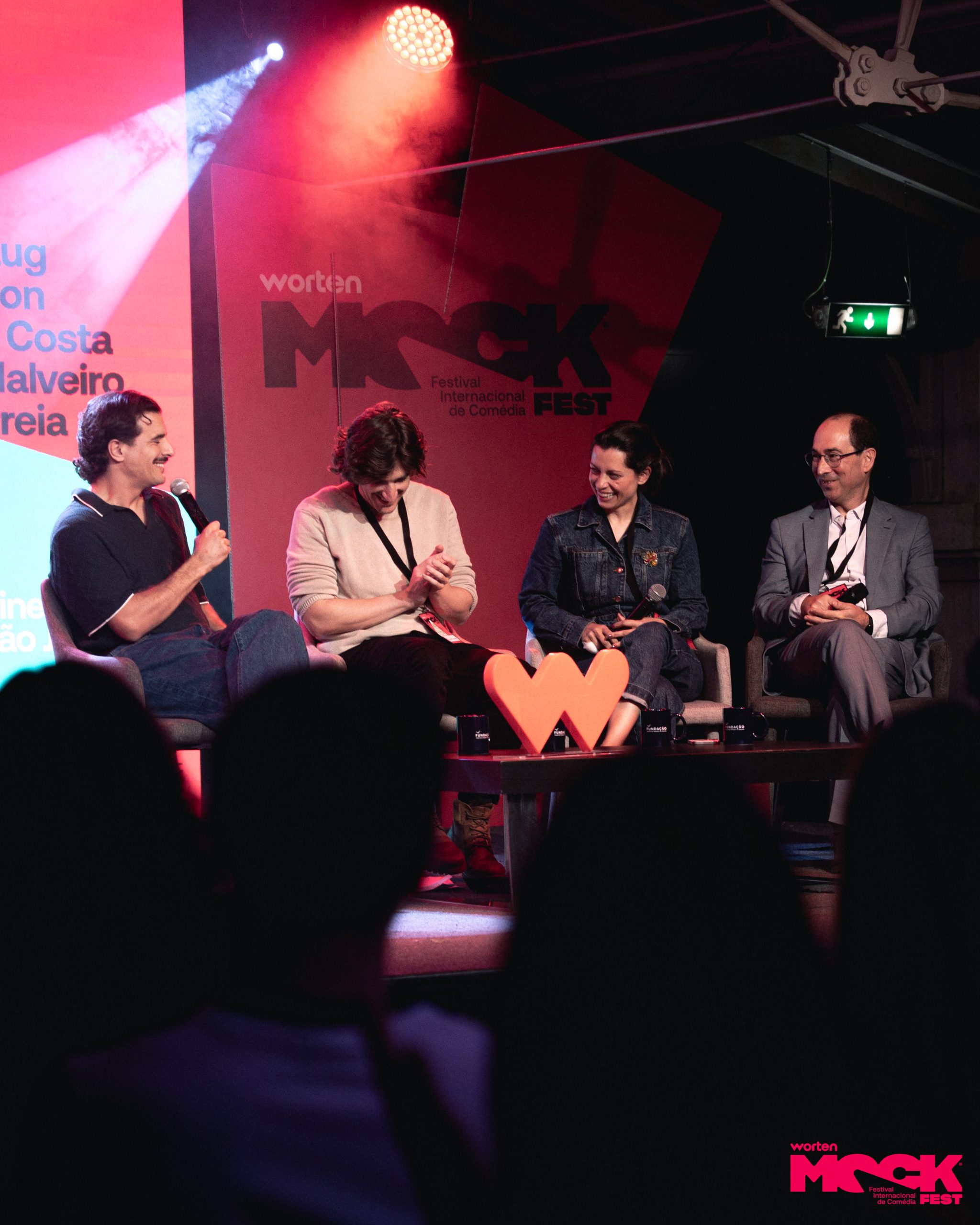Painel de comediantes e convidados sentados no palco durante uma conversa no Worten Mock Fest, com expressões descontraídas e sorridentes.
