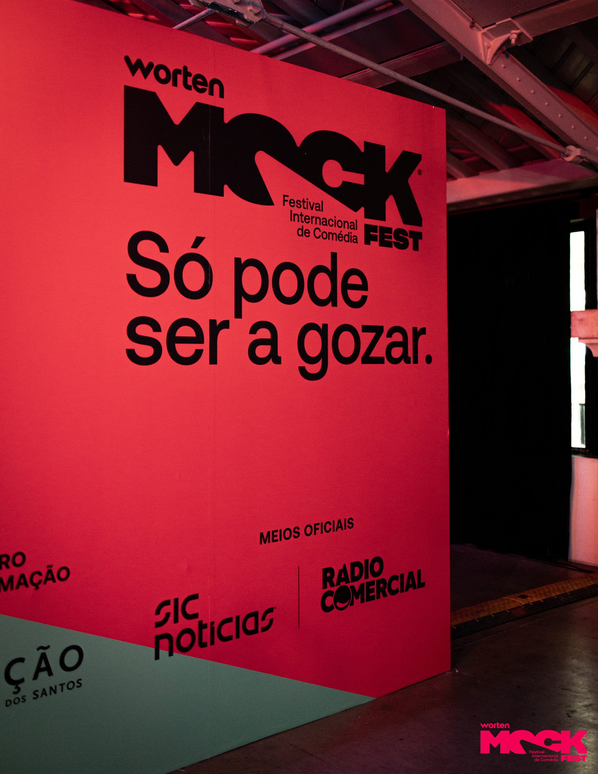 Parede rosa com o logotipo do Worten Mock Fest e o slogan “Só pode ser a gozar”, junto aos logotipos dos meios oficiais SIC Notícias e Rádio Comercial.