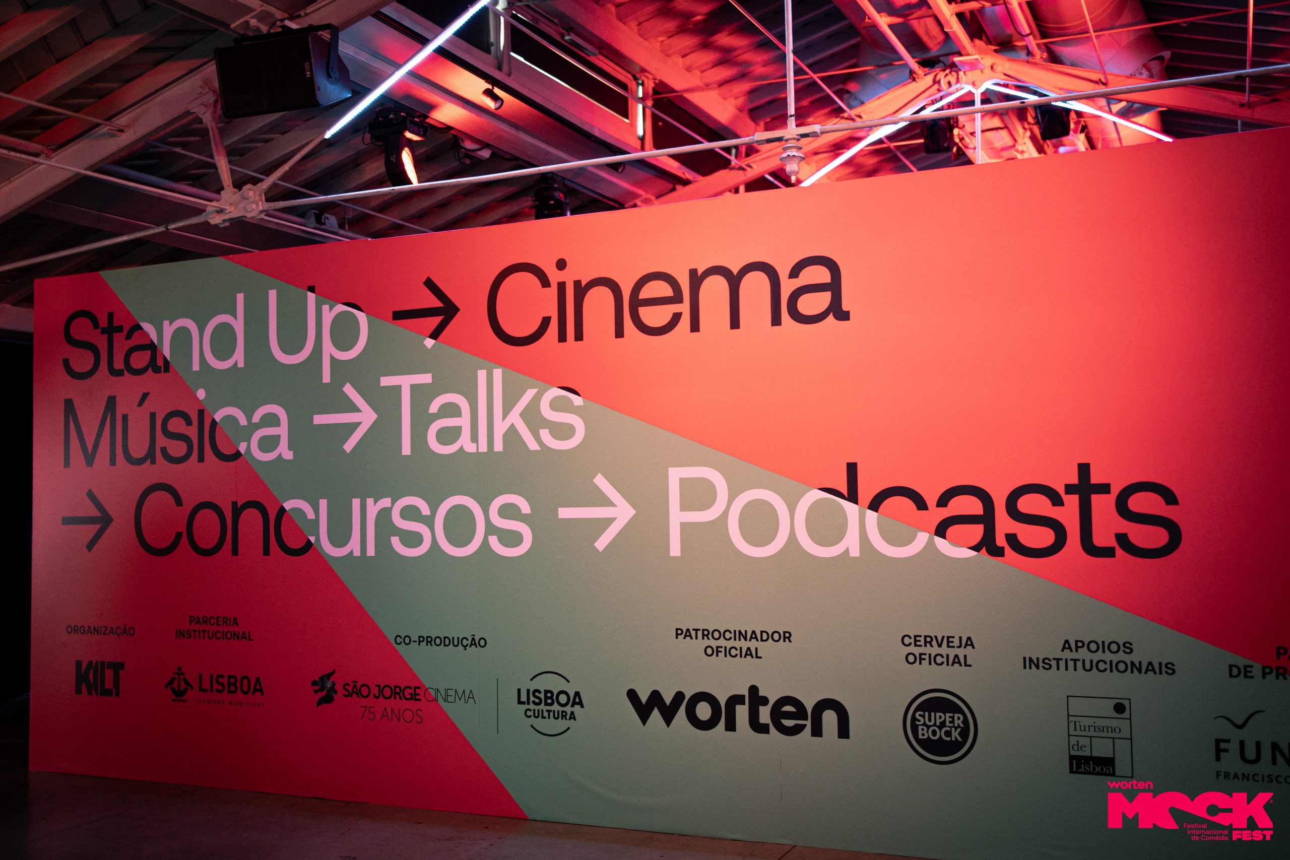 Parede rosa com o logotipo do Worten Mock Fest e o slogan “Só pode ser a gozar”, junto aos logotipos dos meios oficiais SIC Notícias e Rádio Comercial.
