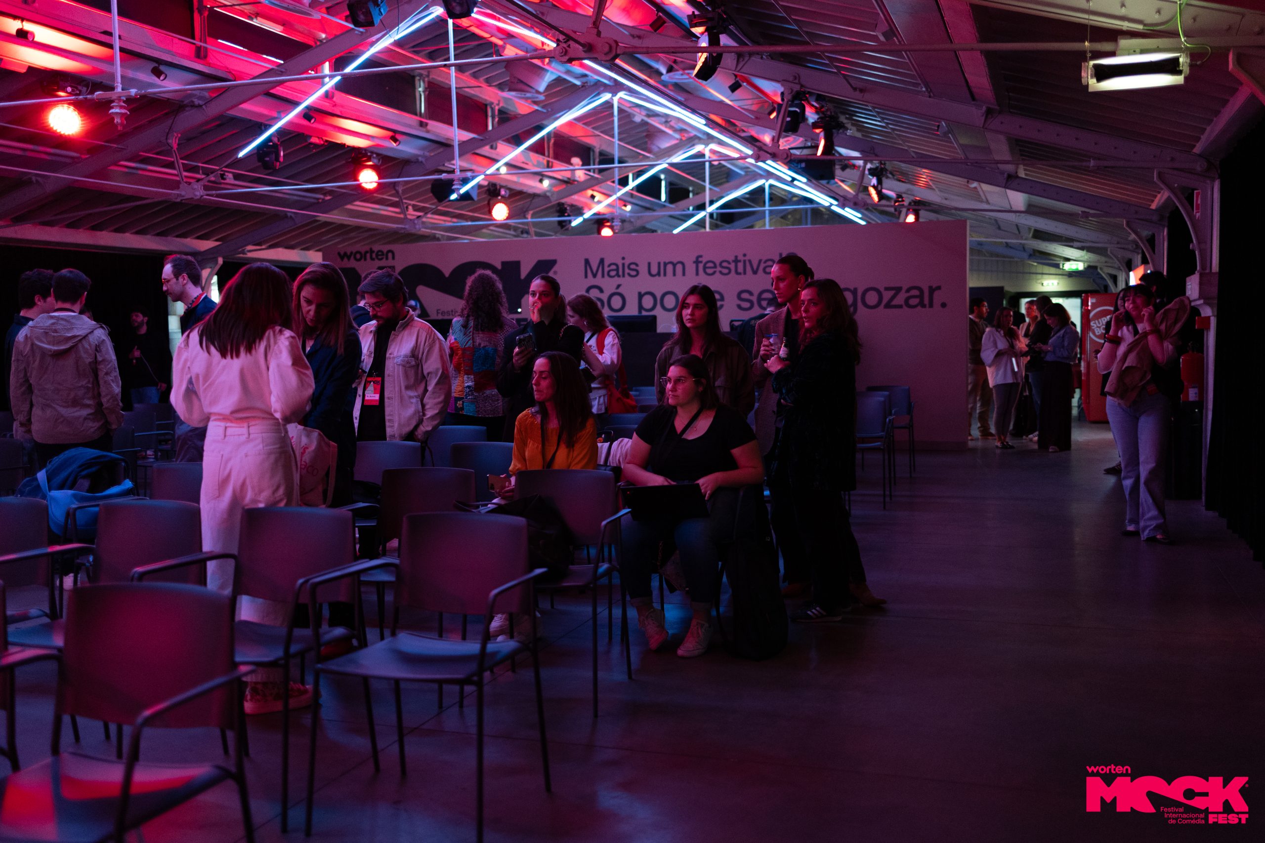 Público circulando e conversando após uma palestra no Worten Mock Fest. Ambiente descontraído, iluminação colorida e clima de festival. Ao fundo, o slogan: “Mais um festival? Só pode ser a gozar.