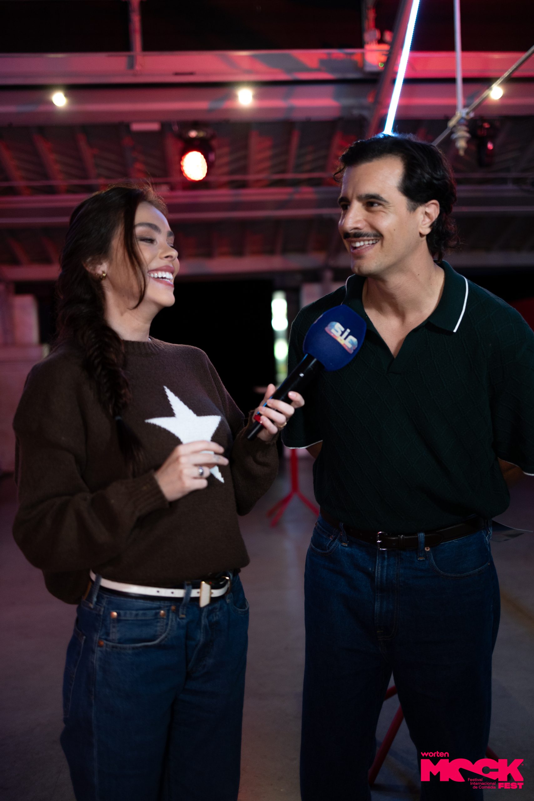 Repórter do canal SIC entrevistando um dos comediantes do Mock Fest, ambos sorrindo durante a gravação no recinto do festival.