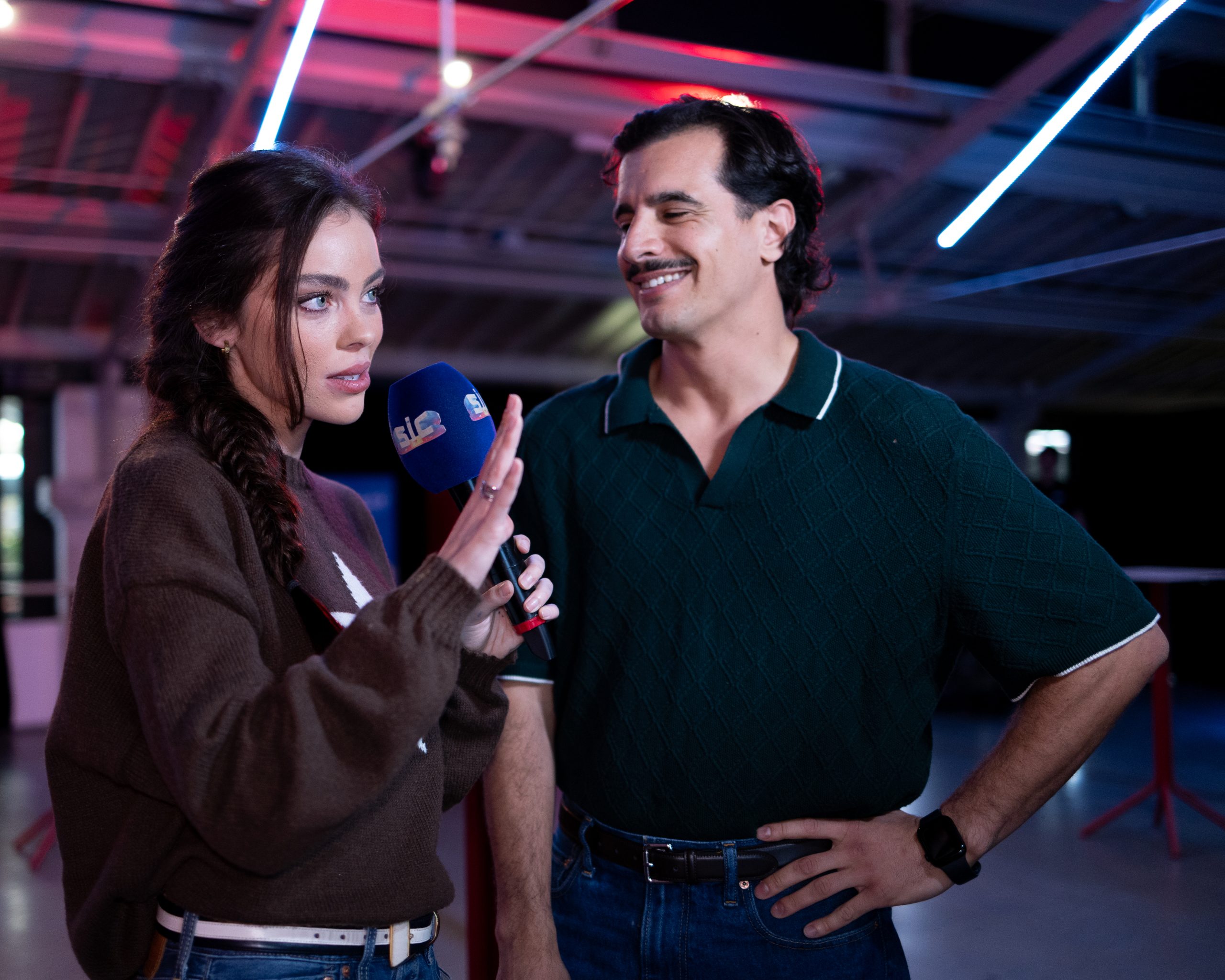 A female presenter holds a blue microphone while engaging with a male interviewee in a modern studio setting.
