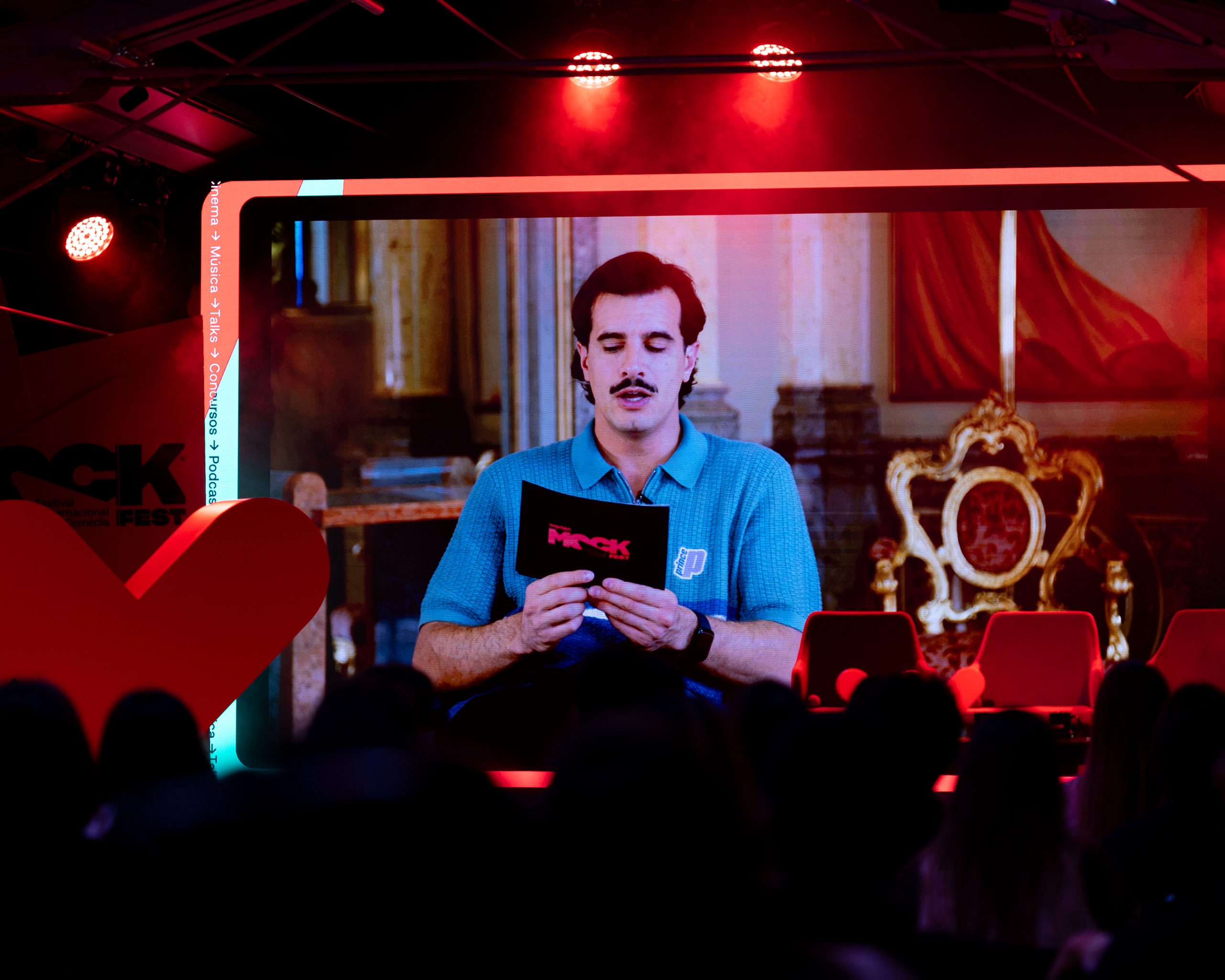 Homem de bigode e camisa azul aparece em um telão segurando um cartão com o logo do Mock Fest. Ao fundo, cenário com cadeiras douradas e cortinas vermelhas, enquanto o público assiste.