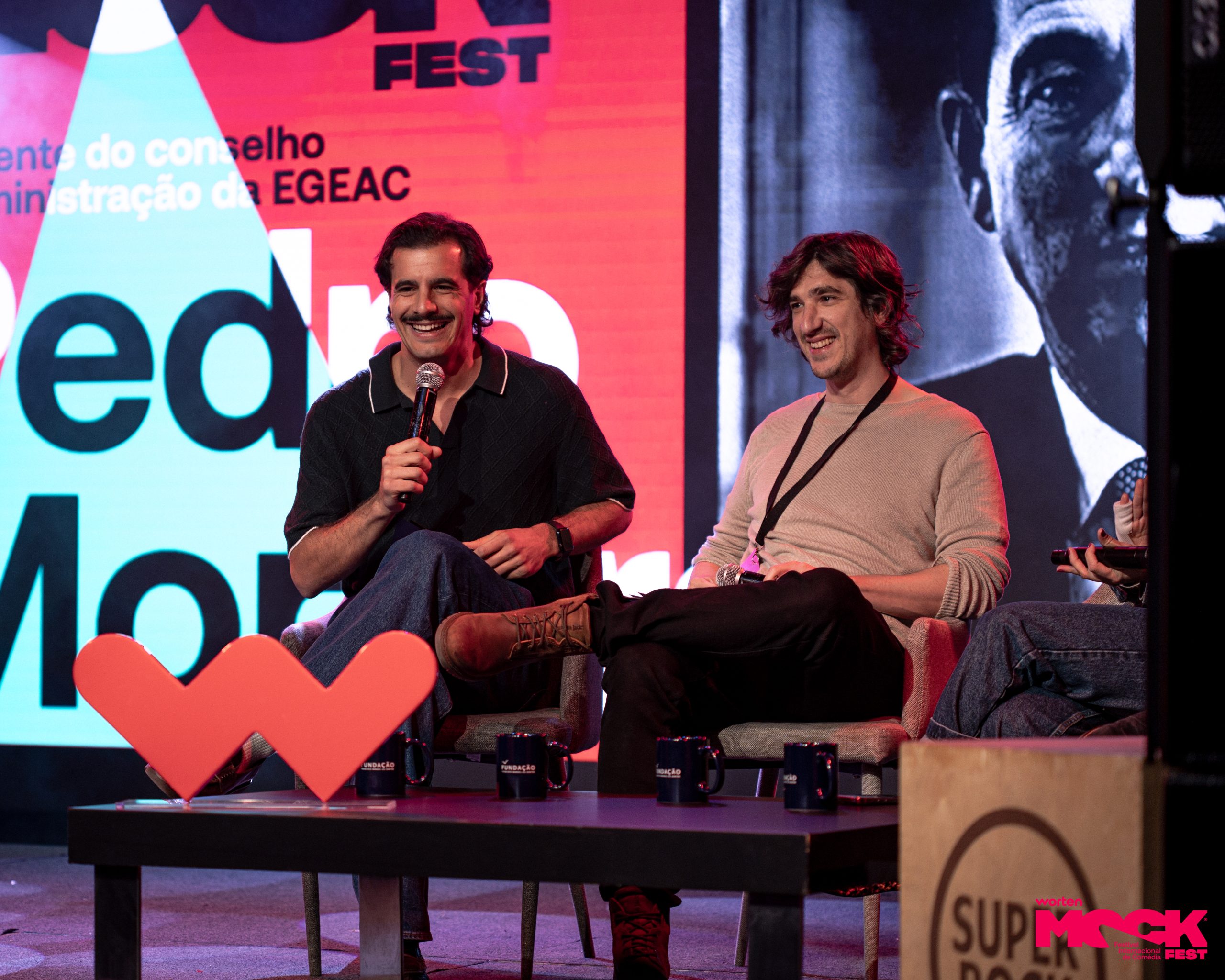 Painel de discussão no Worten Mock Fest. Três pessoas conversam, duas segurando microfones, com expressão atenta e envolvida. O fundo traz o logo do evento e luzes coloridas