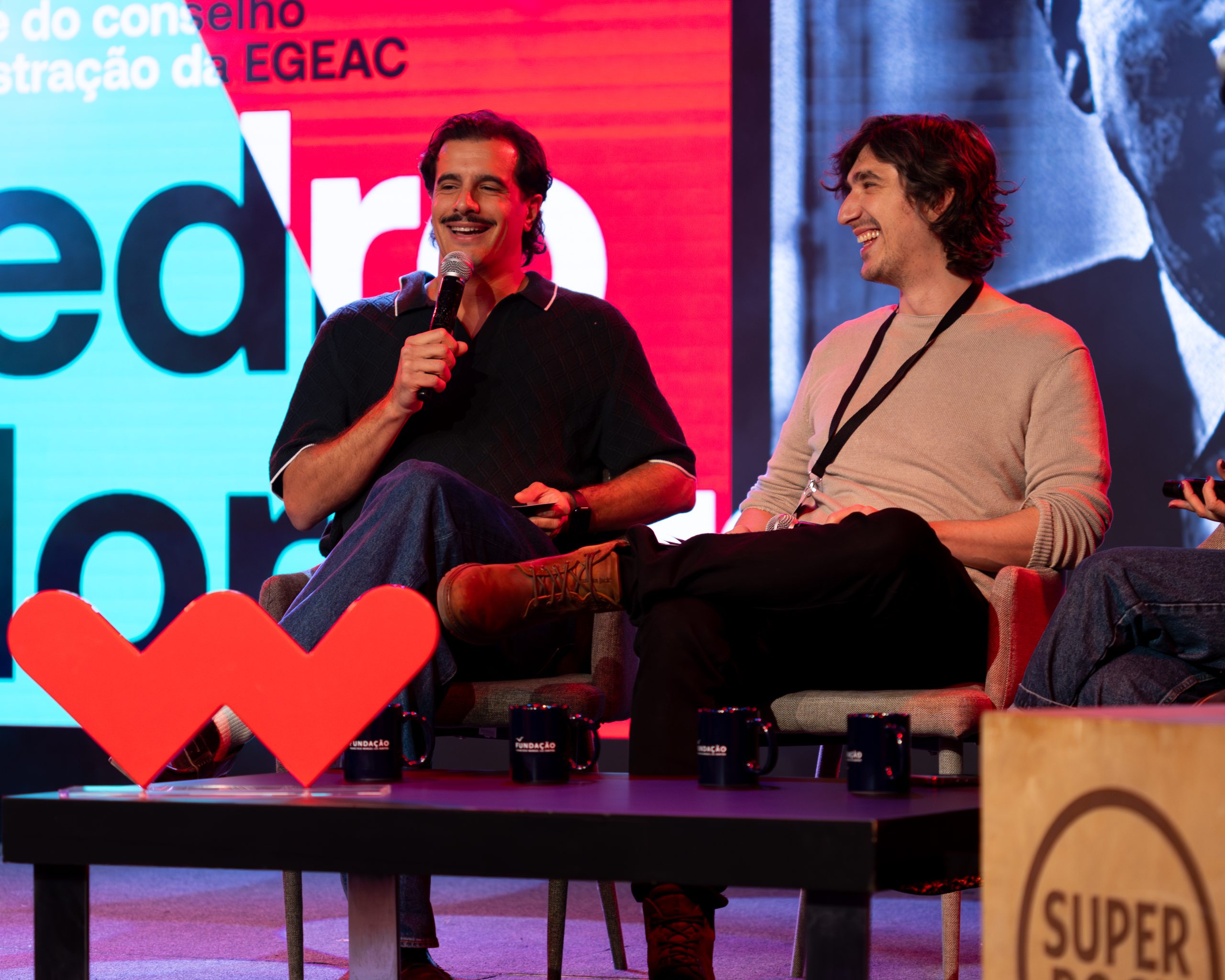 Comediantes no palco do Worten Mock Fest em um painel animado, sorrindo e conversando diante de uma plateia, com um grande logotipo vermelho em destaque à frente.