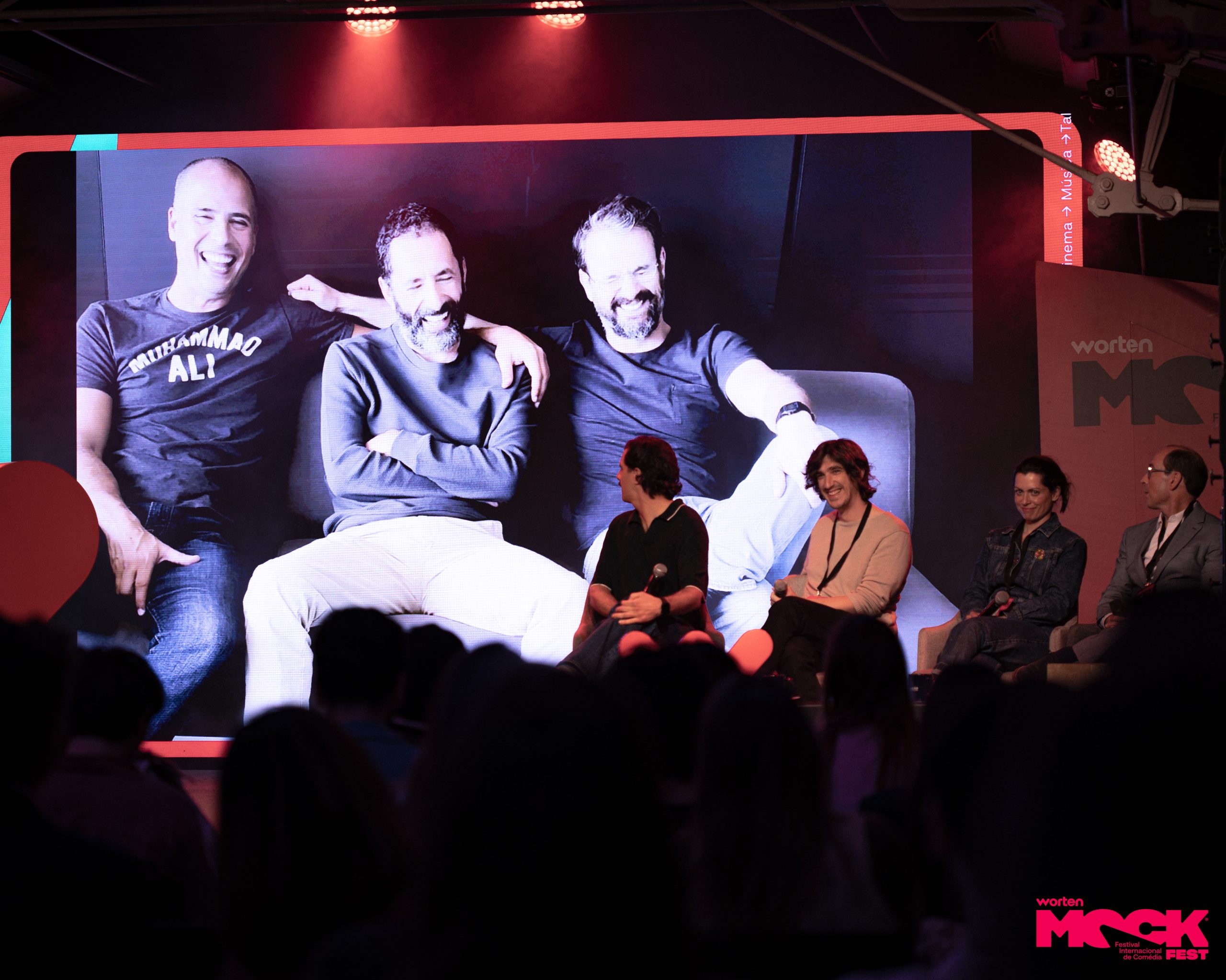 Painel do Mock Fest com convidados sentados no palco assistindo a uma grande tela exibindo uma foto em preto e branco de três homens sorrindo, em ambiente descontraído e público ao fundo.