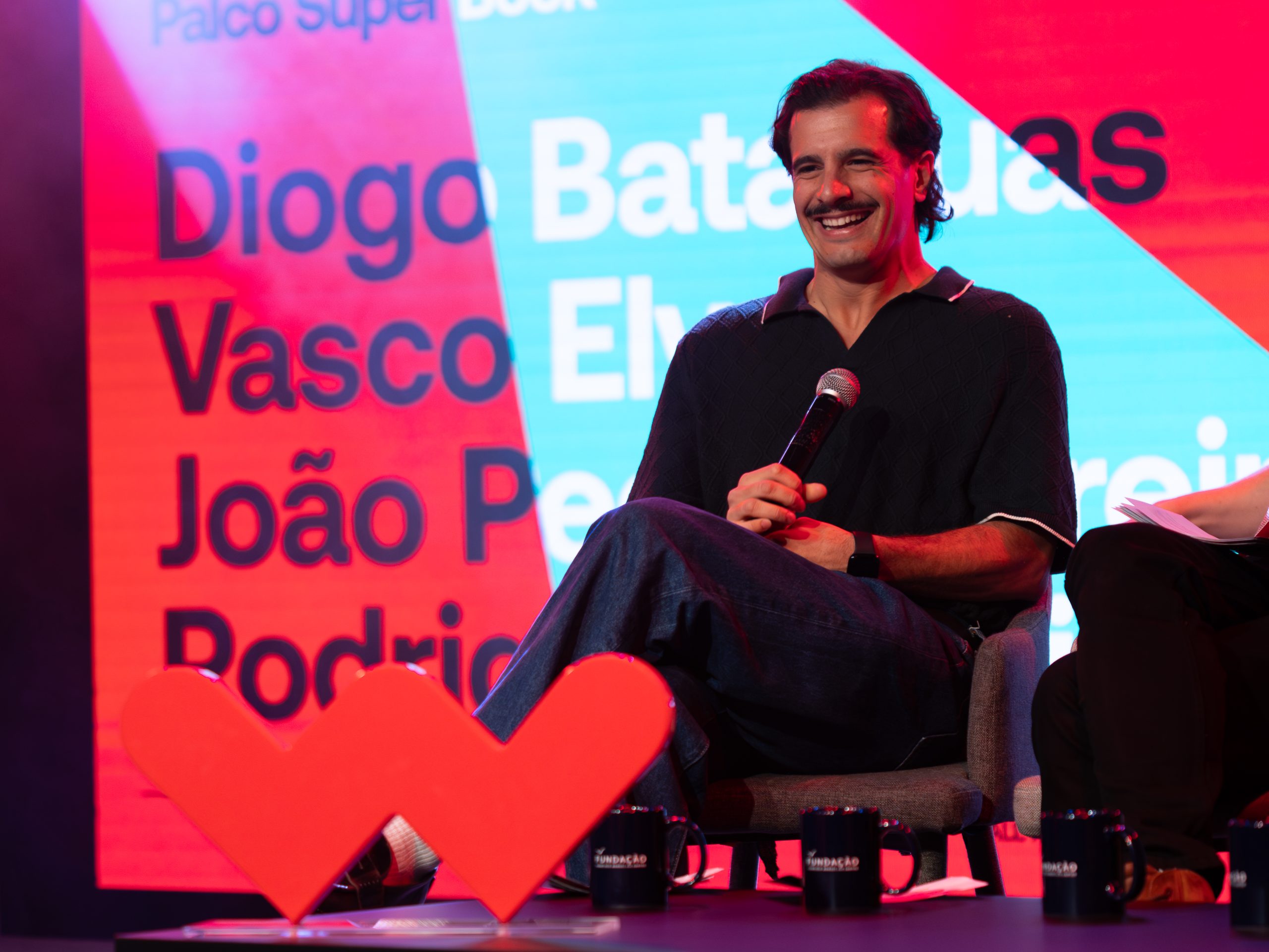 Humorista sendo entrevistado e fotografado no Worten Mock Fest em Lisboa, sorrindo diante do painel do festival de comédia.