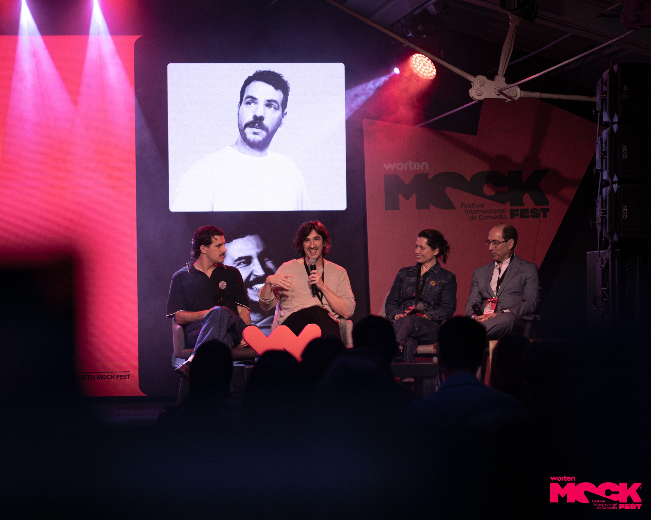 Painel de humoristas no palco do Worten Mock Fest em Lisboa, com público assistindo, projeção de retrato de artista e iluminação de festival de comédia ao vivo.