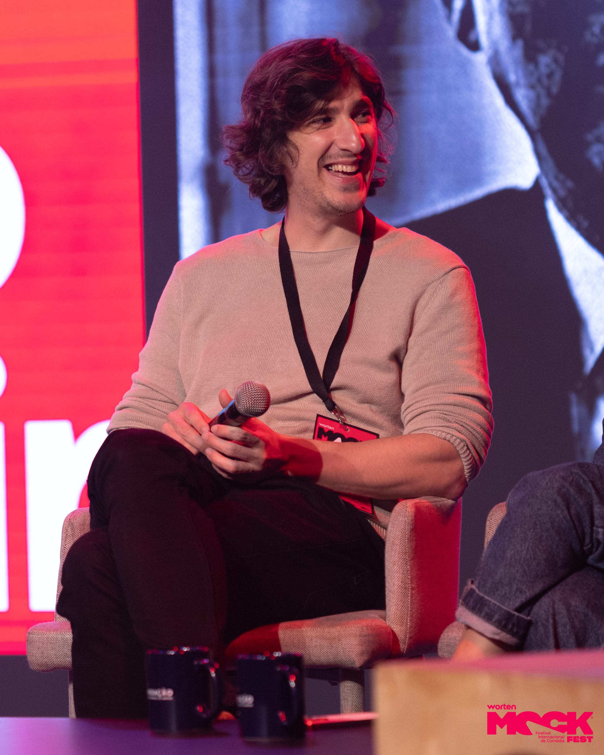 Comediante sentado no palco do Worten Mock Fest, sorrindo e segurando um microfone durante um painel do festival.