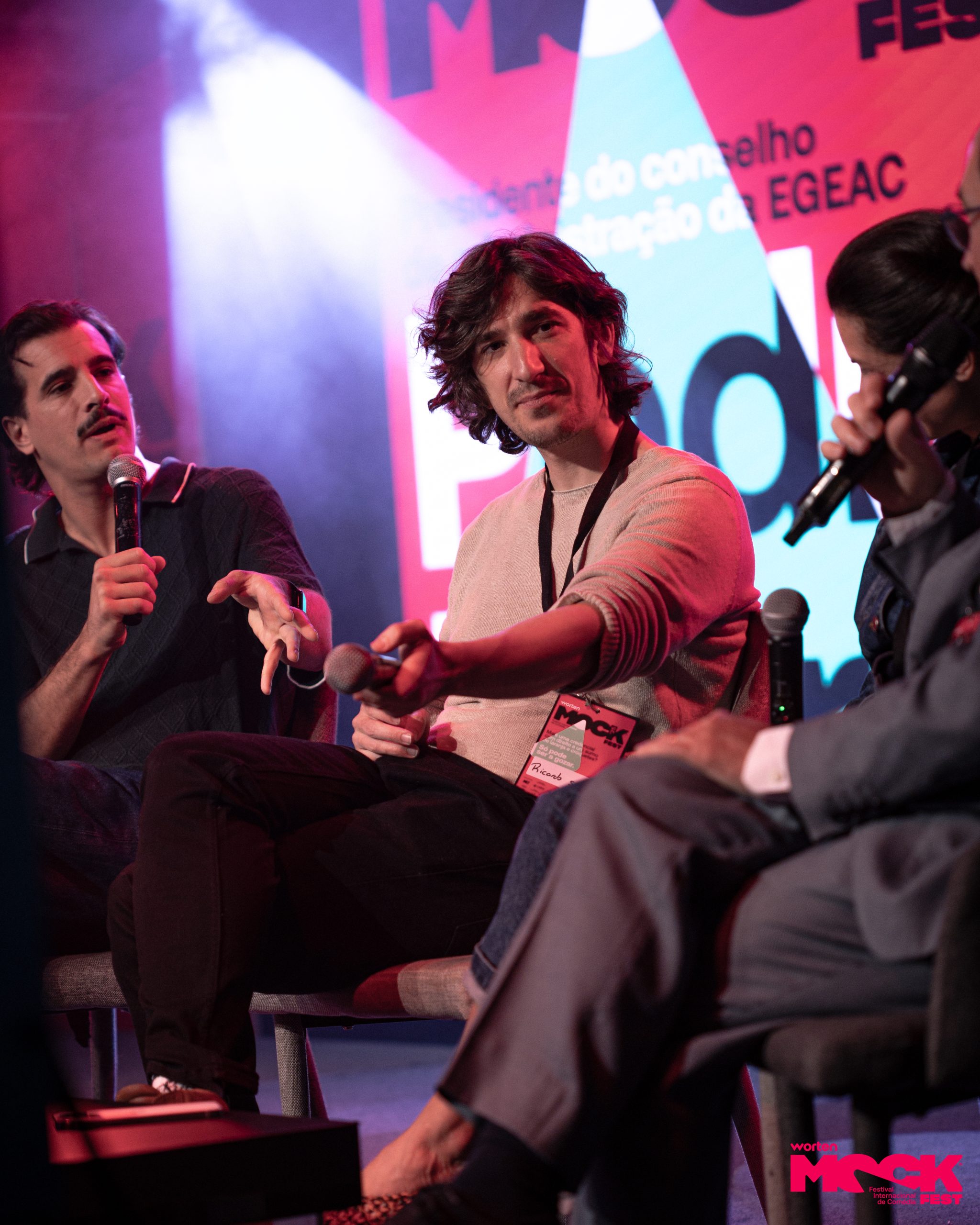 Painel de discussão no Worten Mock Fest. Três pessoas conversam, duas segurando microfones, com expressão atenta e envolvida. O fundo traz o logo do evento e luzes coloridas, criando um ambiente descontraído e criativo.