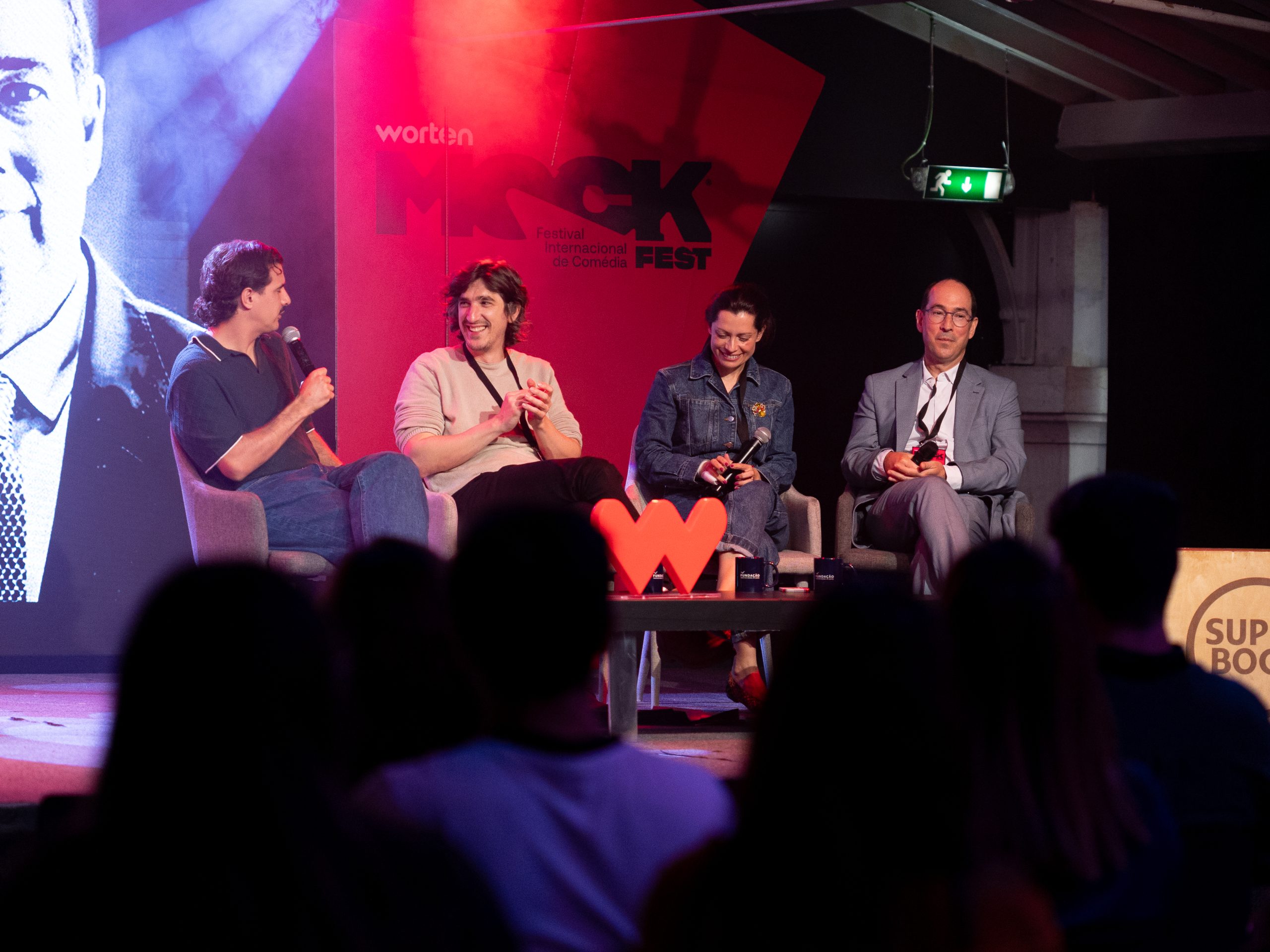 Painel de convidados no palco do Worten Mock Fest, com quatro pessoas sentadas em cadeiras discutindo, microfones nas mãos, público em primeiro plano e logotipo do festival ao fundo.