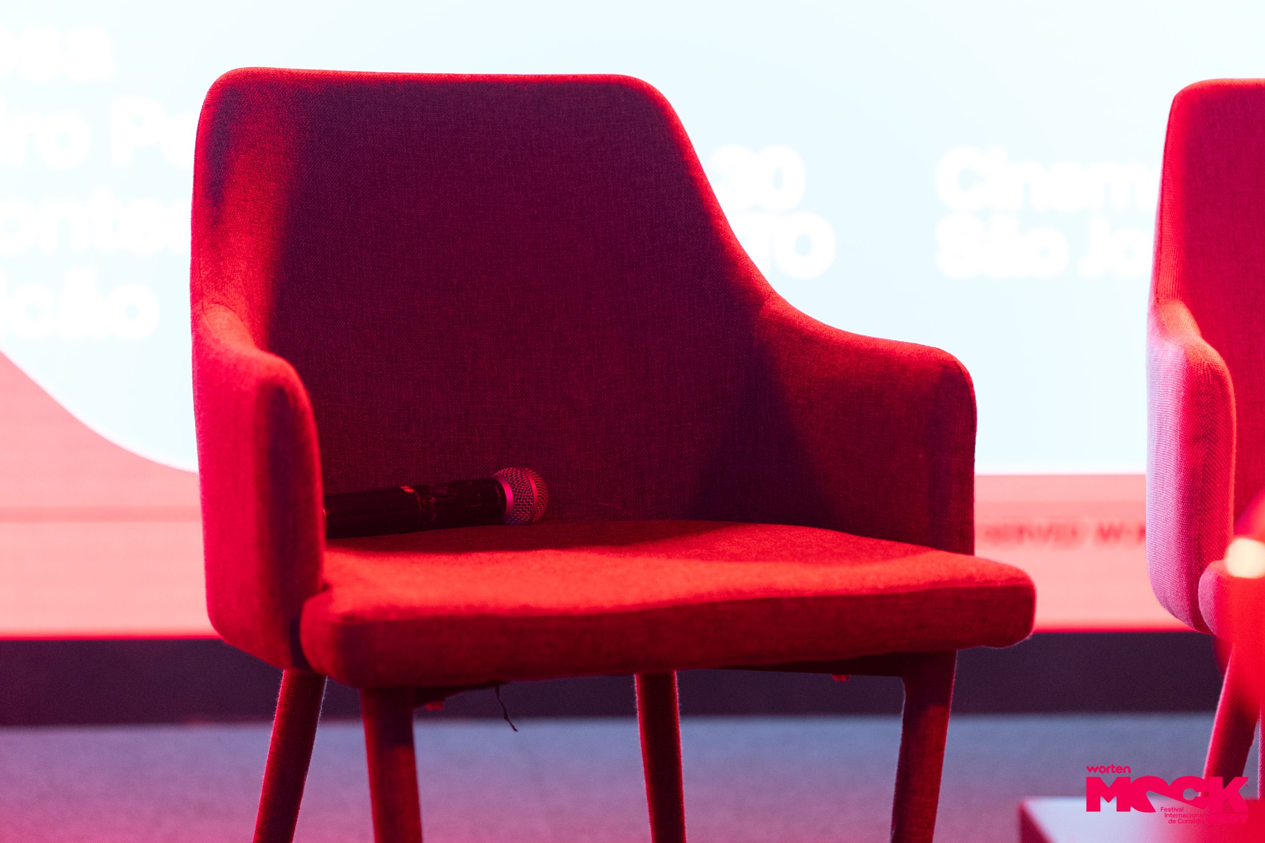 Uma cadeira vermelha vazia no palco com um microfone em cima do assento. Ao fundo, um ecrã claro com texto desfocado e, no canto inferior direito, o logotipo do Worten Mock Fest.