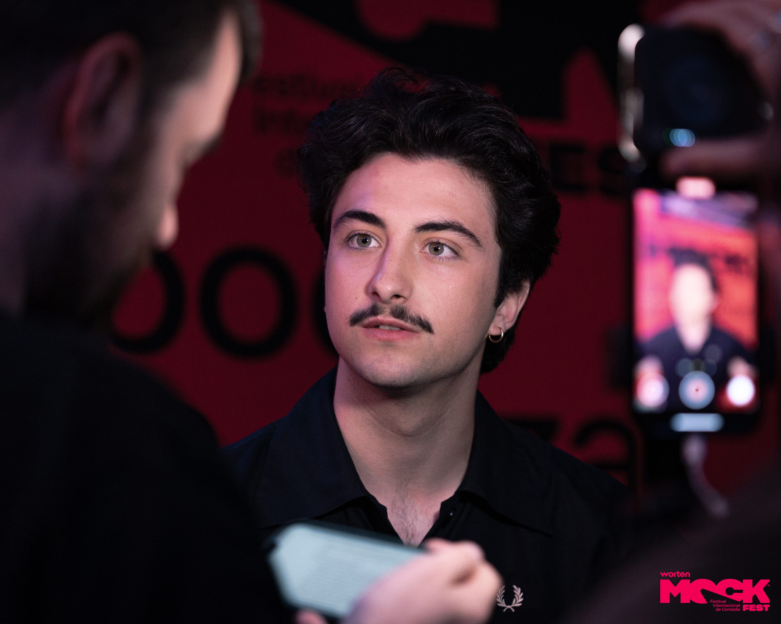 Humorista sendo entrevistado e fotografado no Worten Mock Fest em Lisboa, sorrindo diante do painel do festival de comédia.