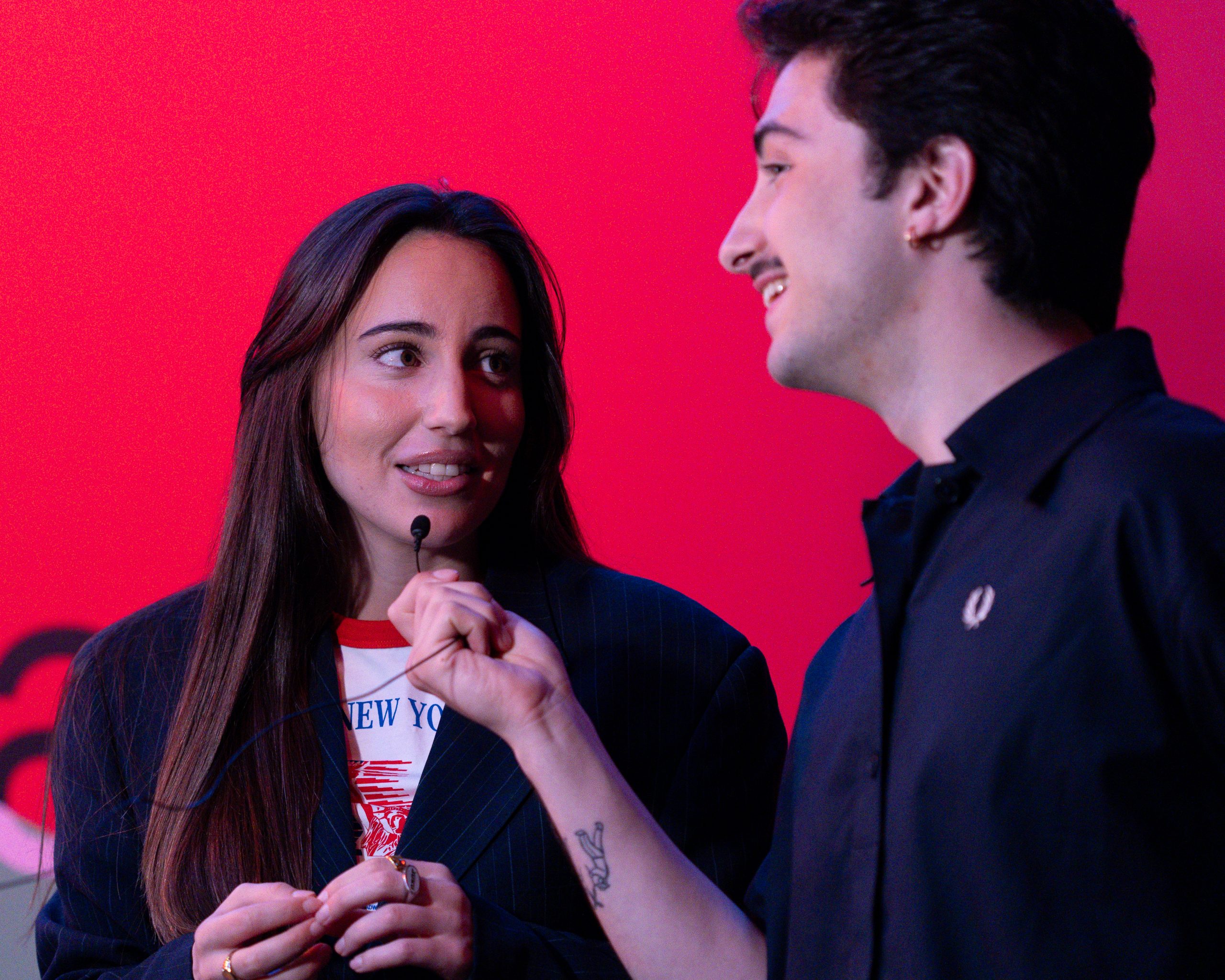 Dois apresentadores do Mock Fest, sorrindo no palco durante uma entrevista, com microfone na mão e fundo vermelho com o logo do festival.