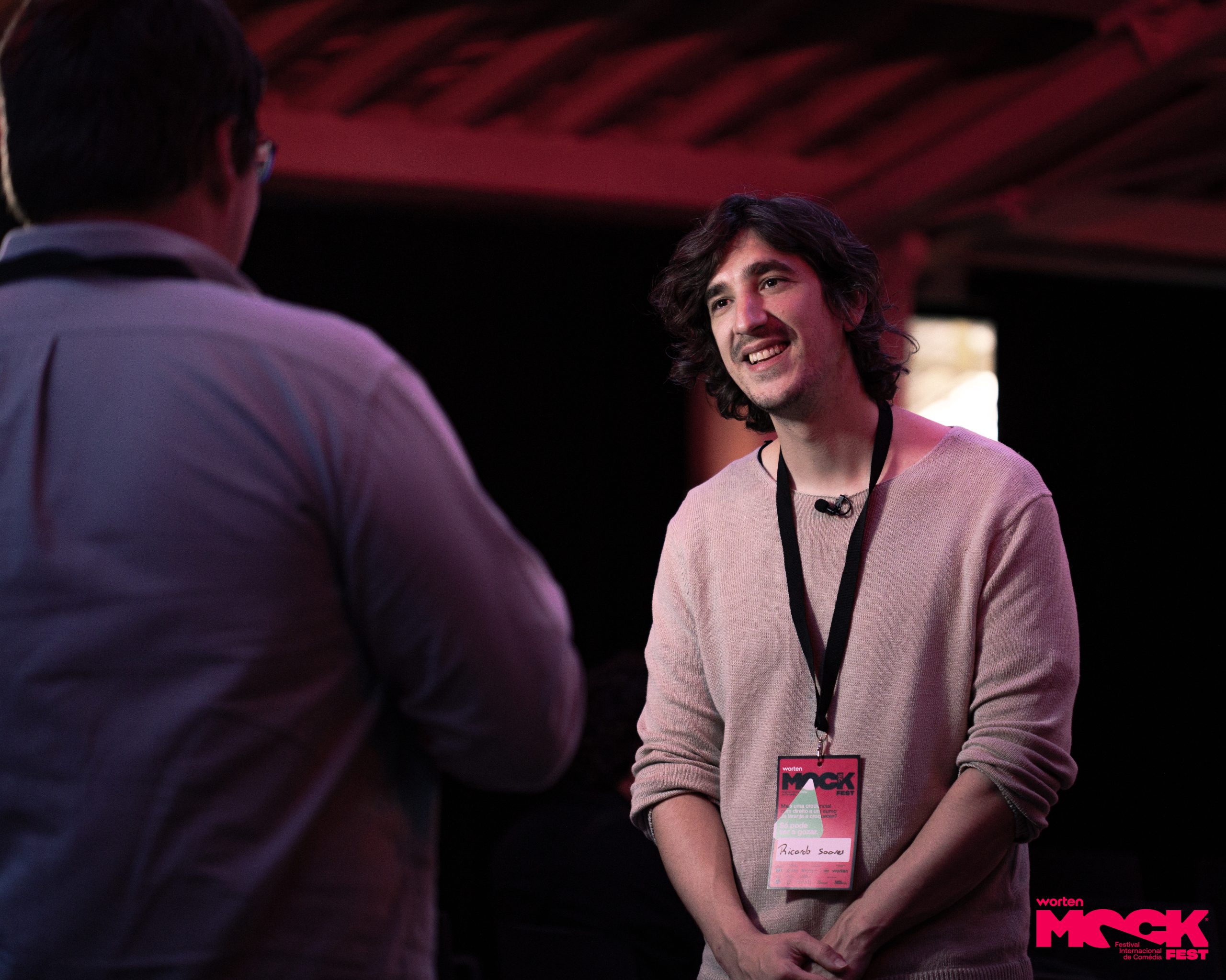 Ricardo, participante do Mock Fest, usando crachá e microfone de lapela, conversa com outra pessoa durante o festival de comédia.