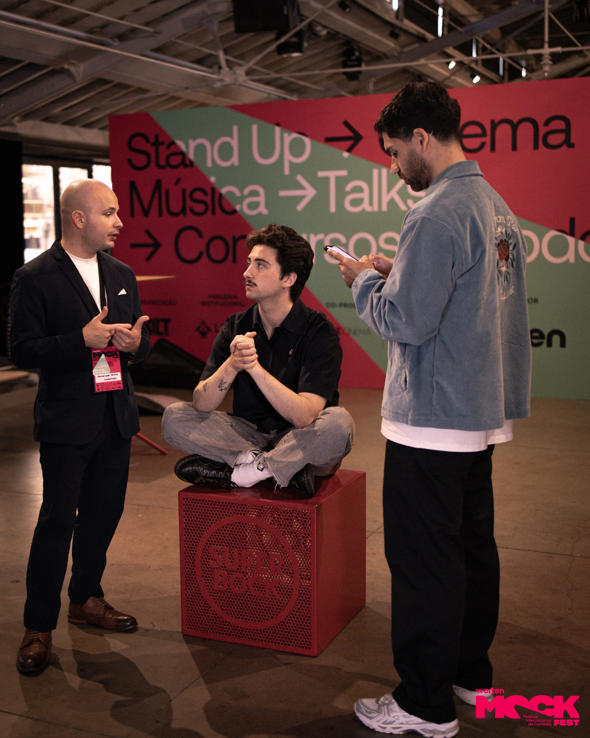 Participantes e artistas conversando na área de convivência do Worten Mock Fest, com painel de fundo destacando stand-up, música e talks no festival de comédia em Lisboa.